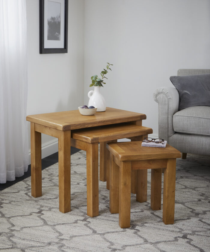 lifestyle image of a nest of three oak tables with a vase and decorative bowl on top, set in a sitting room on a rug next to a grey couch.